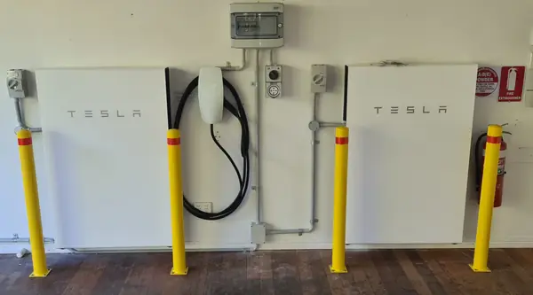 Two Tesla Powerwall batteries and a wall connector mounted on a wall behind safety bollards